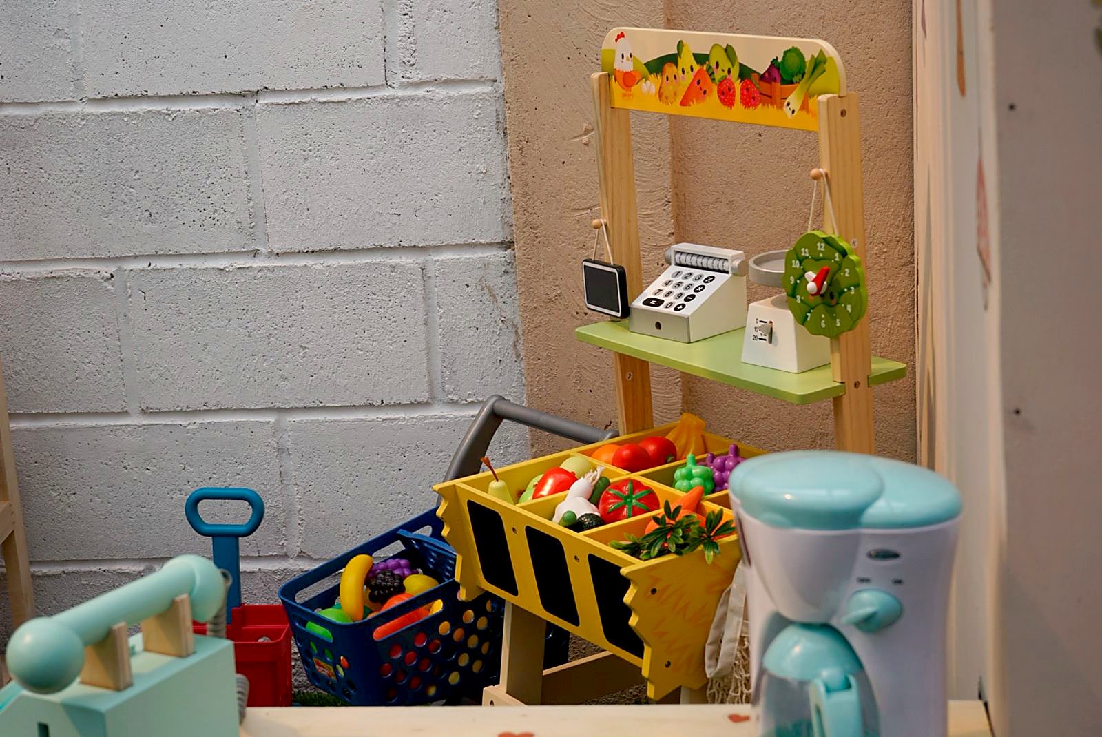 Detalle del mercadito de juguete: carrito de compras con frutas y verduras, caja registradora y cafetera